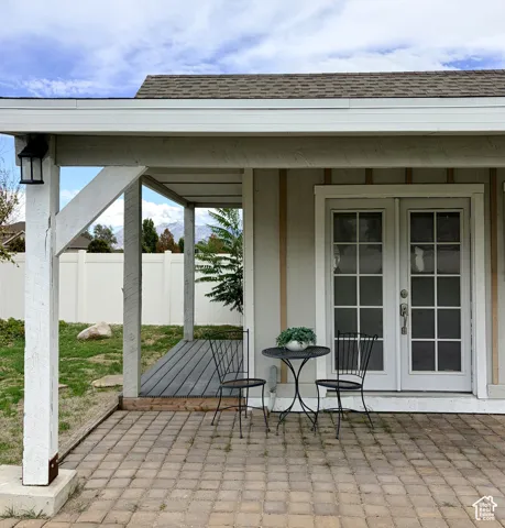 Paved patio area, covered porch with exposed wood beams, exterior wall lantern, double glass-paneled doors, and a partial privacy fence