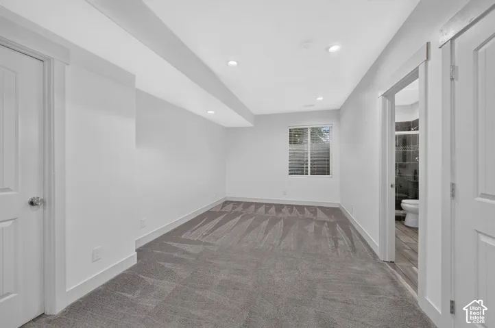 Recessed ceiling lighting, window with horizontal blinds, grey patterned carpet, white interior doors, and wood-finish flooring in the adjacent room