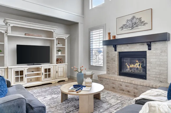 Two-story ceiling, brick fireplace with a dark wood mantel, built-in media center with shelving, plantation shutters, and wood-finish flooring