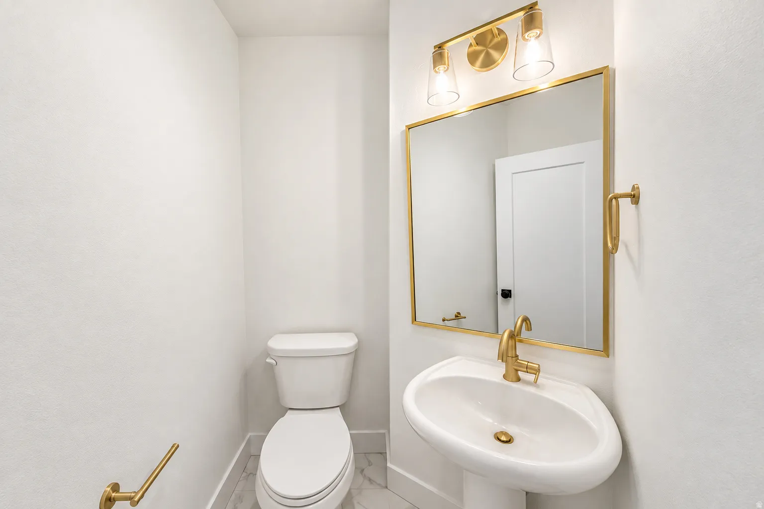 Pedestal sink with brass-finish faucet, rectangular mirror with brass-finish frame, brass-finish vanity light fixture, white toilet, and tile flooring