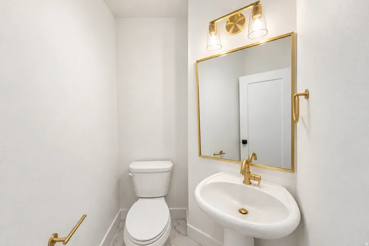 Pedestal sink with brass-finish faucet, rectangular mirror with brass-finish frame, brass-finish vanity light fixture, white toilet, and tile flooring