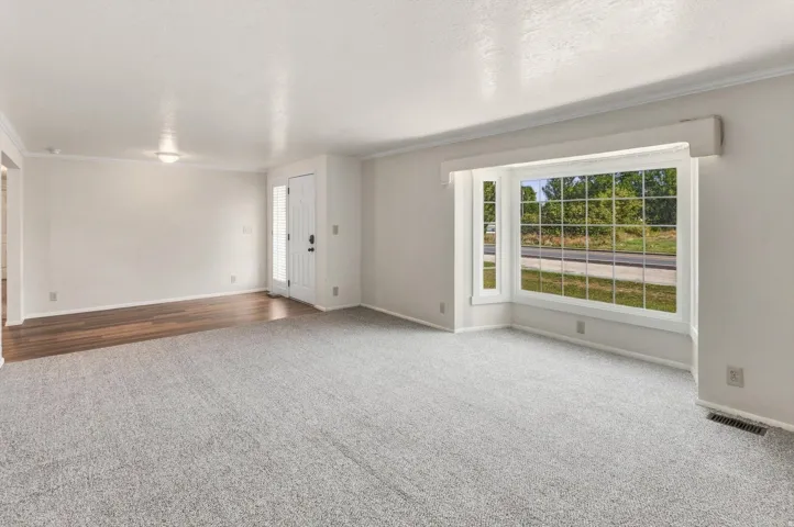 Spacious living area featuring light gray carpeting, a large bay window, white trim, and a white front entry door with a frosted glass panel
