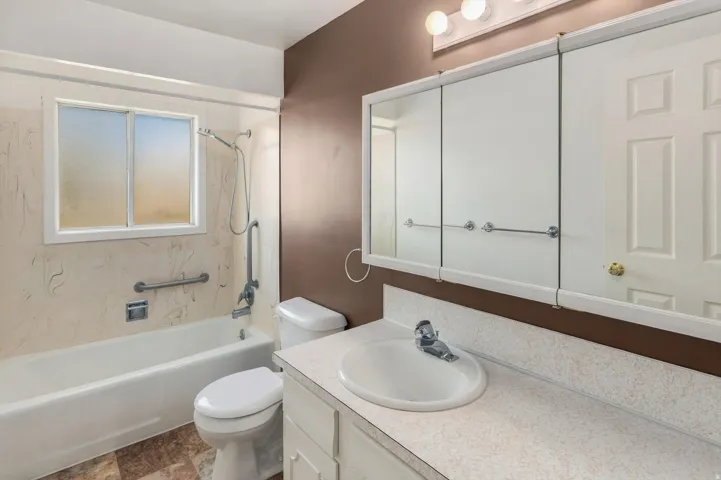 Vanity with integrated sink, three-door mirrored medicine cabinet, white bathtub with grab bars, and a window