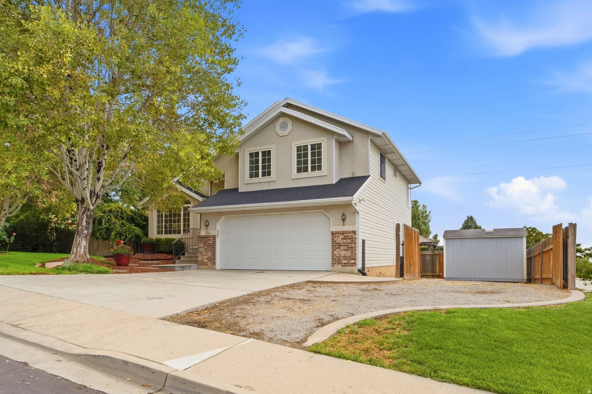 Front view of the home with mature landscaping, large windows, and a two-car garage.  Also showcasing the RV or boat storage to the west side of the home and the detached shed behind double-wide opening gates.