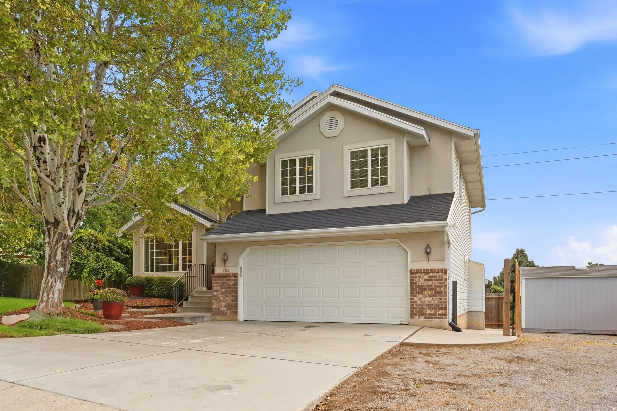 Front view of the home with mature landscaping, large windows, and a two-car garage.  Also showcasing the RV or boat storage to the west side of the home and the detached shed behind double-wide opening gates.