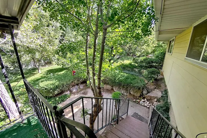 Exterior stairs with black wrought iron railing, leading to a landscaped yard with mature trees and established plantings