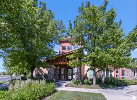 Stone and stucco exterior, prominent entryway with an exposed beam portico, lush landscaping, mature trees, and a clear blue sky
