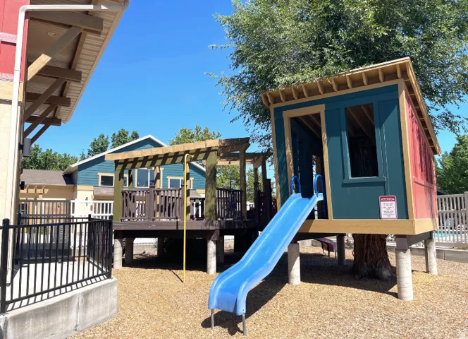 Elevated play structure with an integrated slide, a raised wood-finish deck with pergola, and a black metal railing