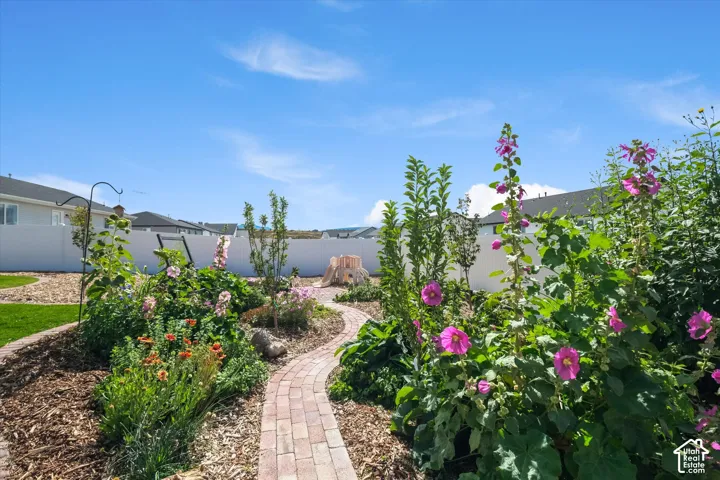 Landscaped garden beds with mature plantings, brick paver pathway, manicured lawn, and a white privacy fence