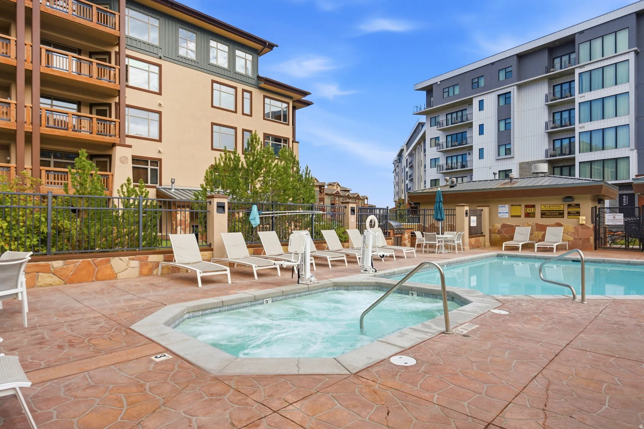 Outdoor pool and spa with stamped concrete surround, surrounded by lounge seating and a stone-clad pool house