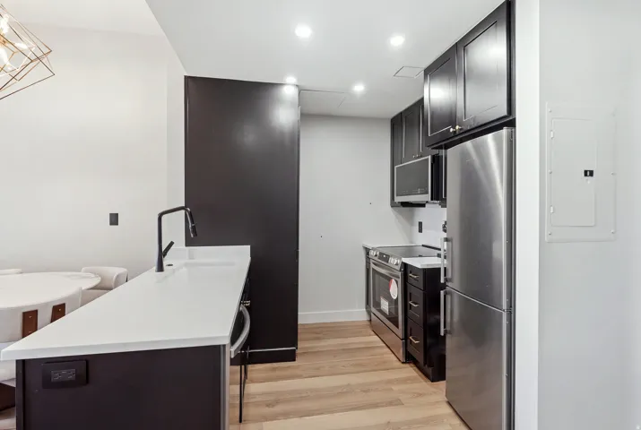White countertop, matte black faucet, dark cabinetry, stainless steel appliances, and wood-finish flooring