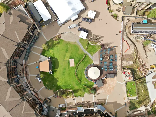 Central green space with meandering pathways, surrounded by paved plaza areas, and multiple commercial structures featuring distinct rooflines