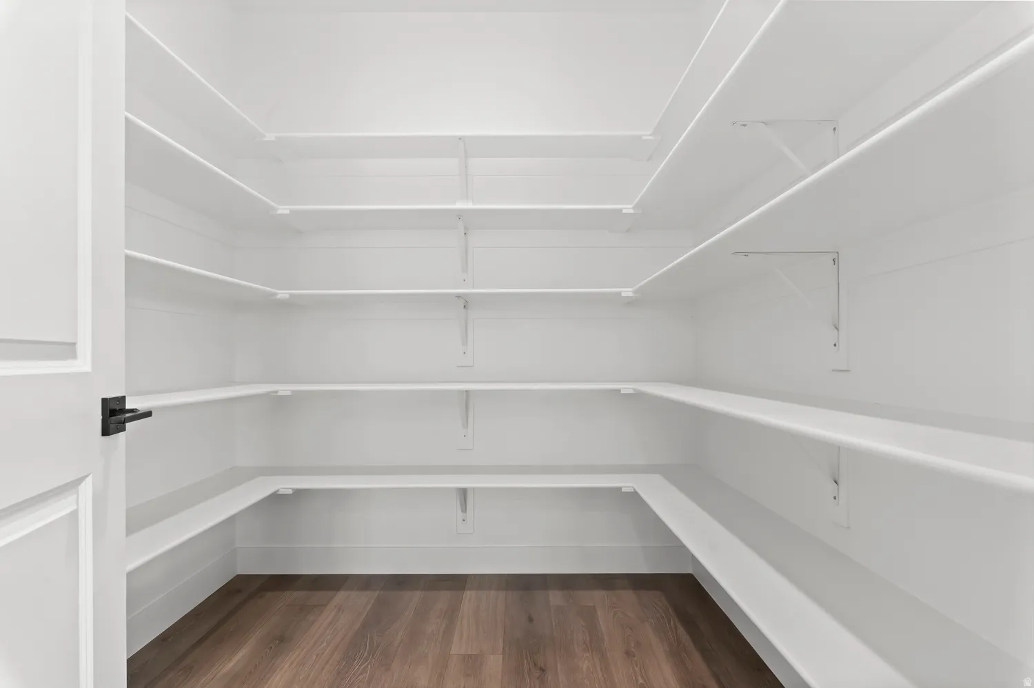 Spacious pantry featuring extensive white shelving, wood-finish flooring, and a modern black door handle