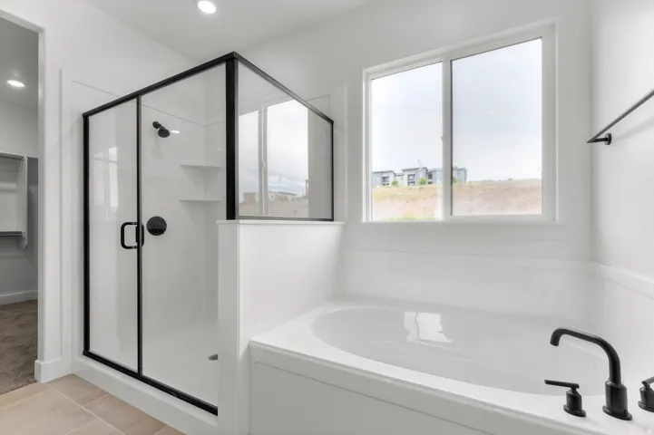 Spacious soaking tub with matte black fixtures, separate shower with black-framed glass enclosure, built-in shower shelving, recessed lighting, and tile flooring