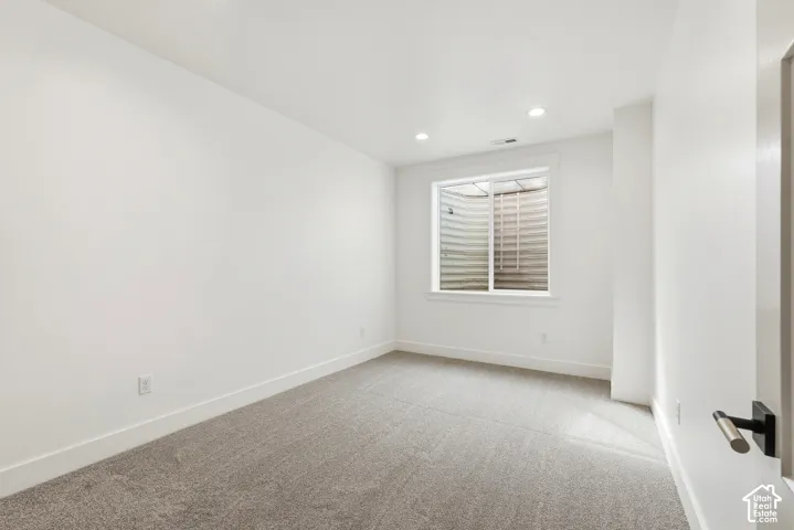 White walls, gray carpeting, recessed lighting, and a single window with a window well