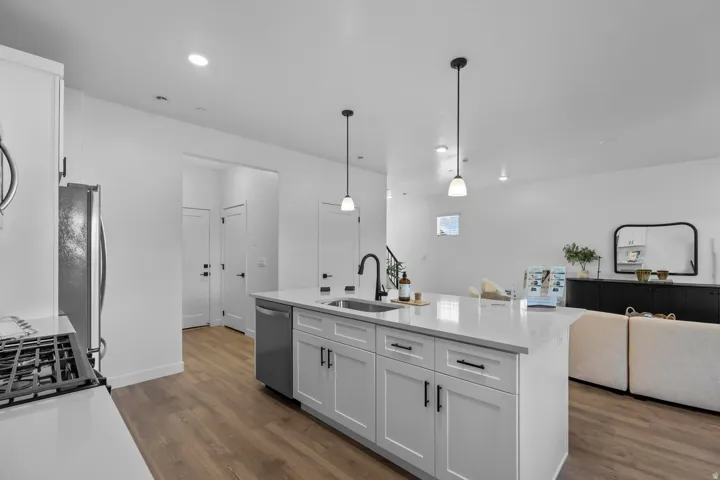 Modern kitchen featuring an island with a sink and pendant lighting, stainless steel appliances, wood-finish flooring, and white cabinetry