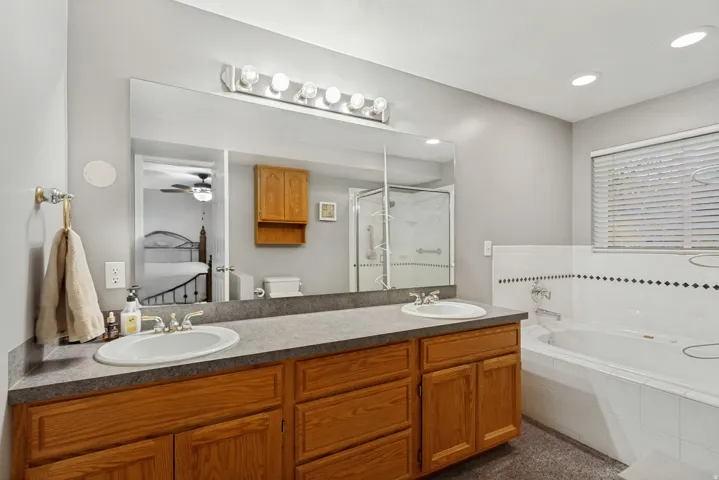 Dual undermount sinks set in a laminate countertop, wood-finish cabinetry, built-in bathtub with tile surround, and a clear glass shower enclosure