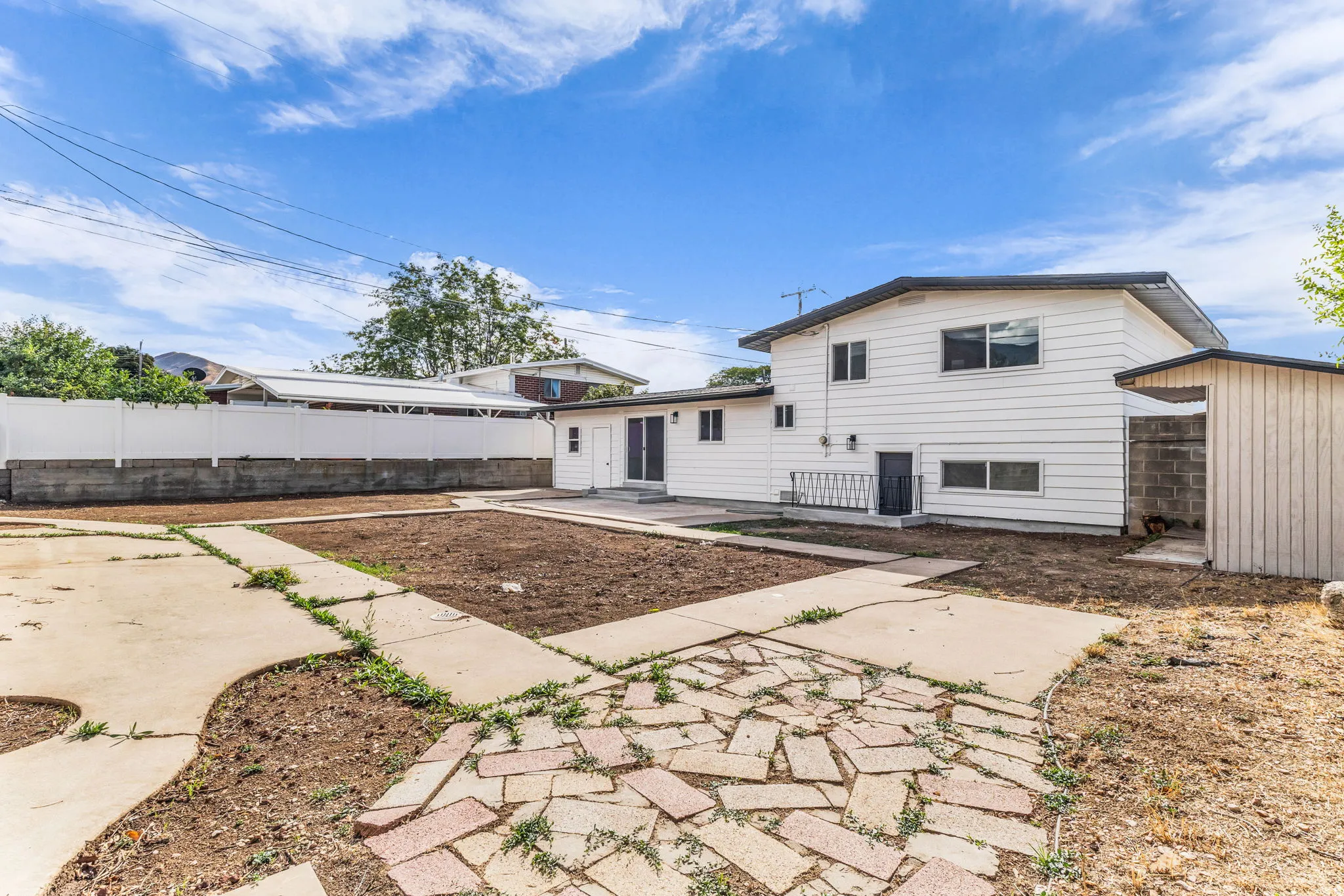 White exterior siding, dark roof, concrete patio, brick paver accents, white privacy fence, and an attached storage shed