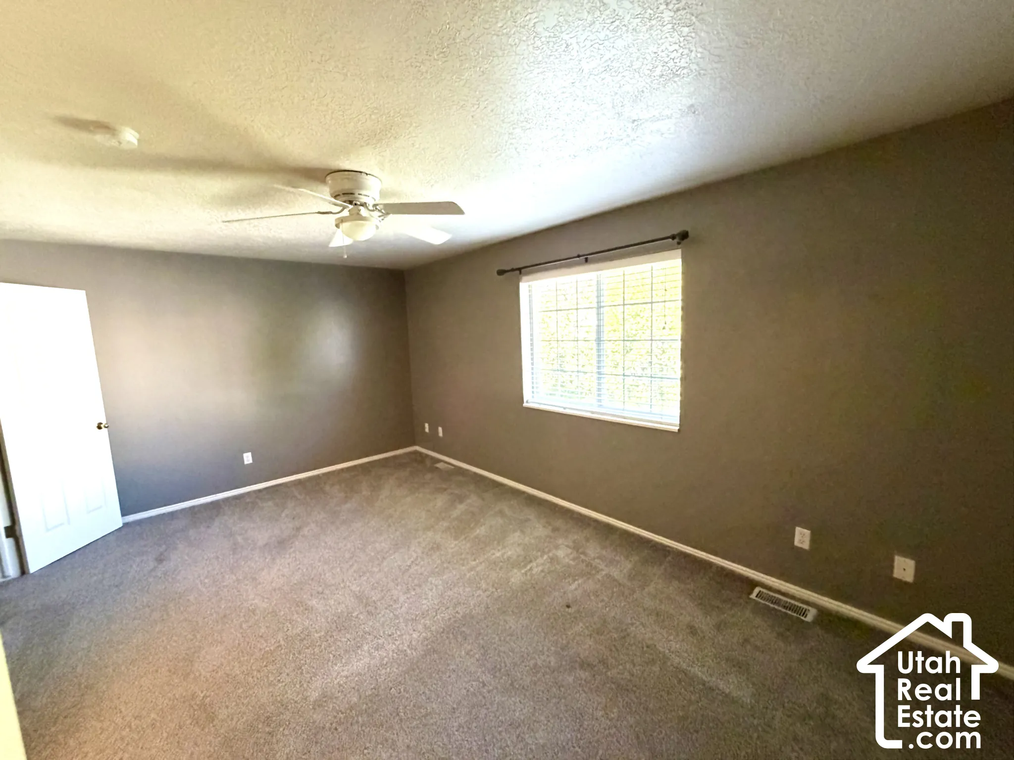 Spacious room featuring neutral carpeting, a ceiling fan with integrated lighting, and a prominent window with blinds