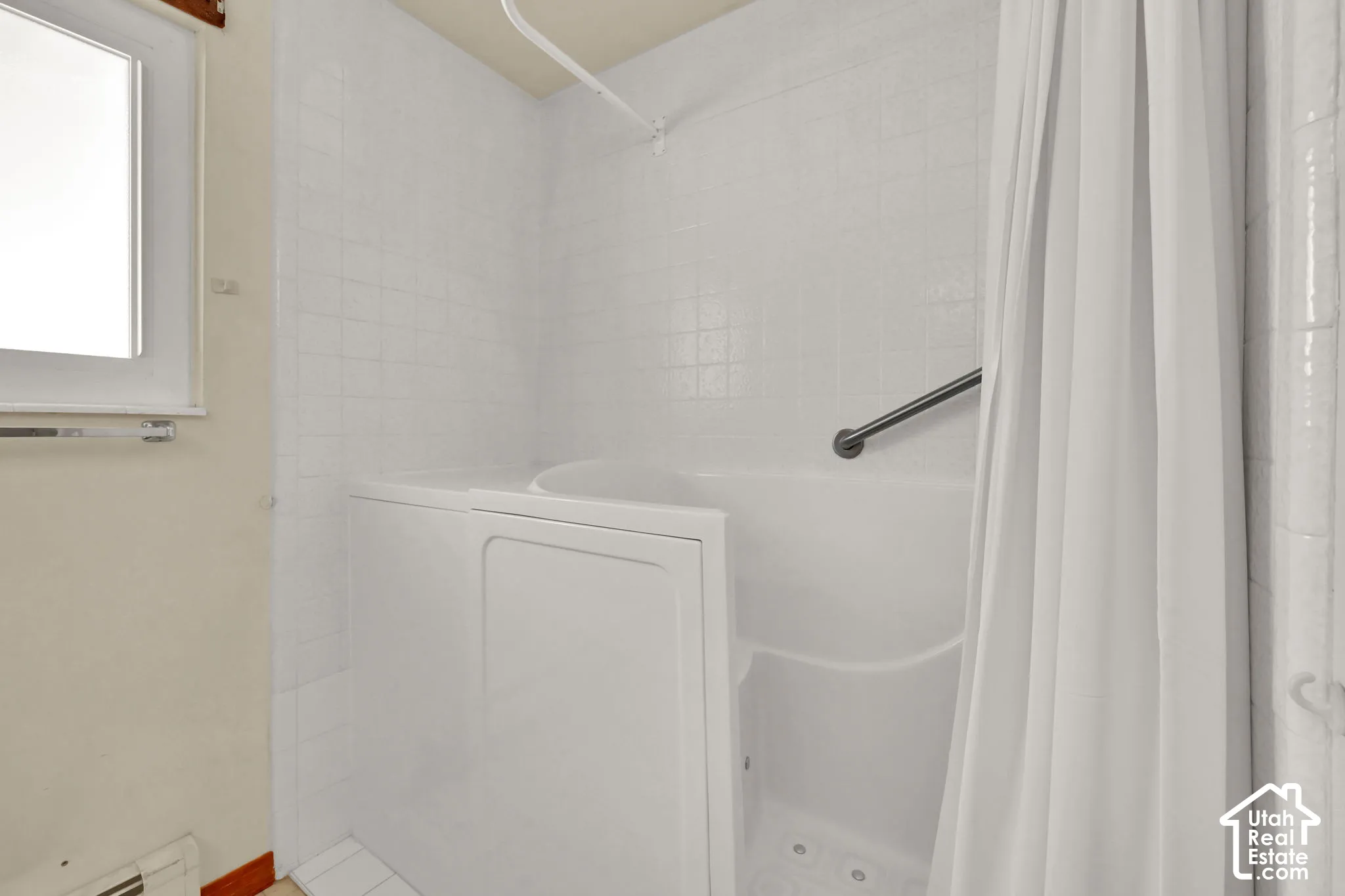 Walk-in tub with a grab bar, white tile surround, and a window providing natural light