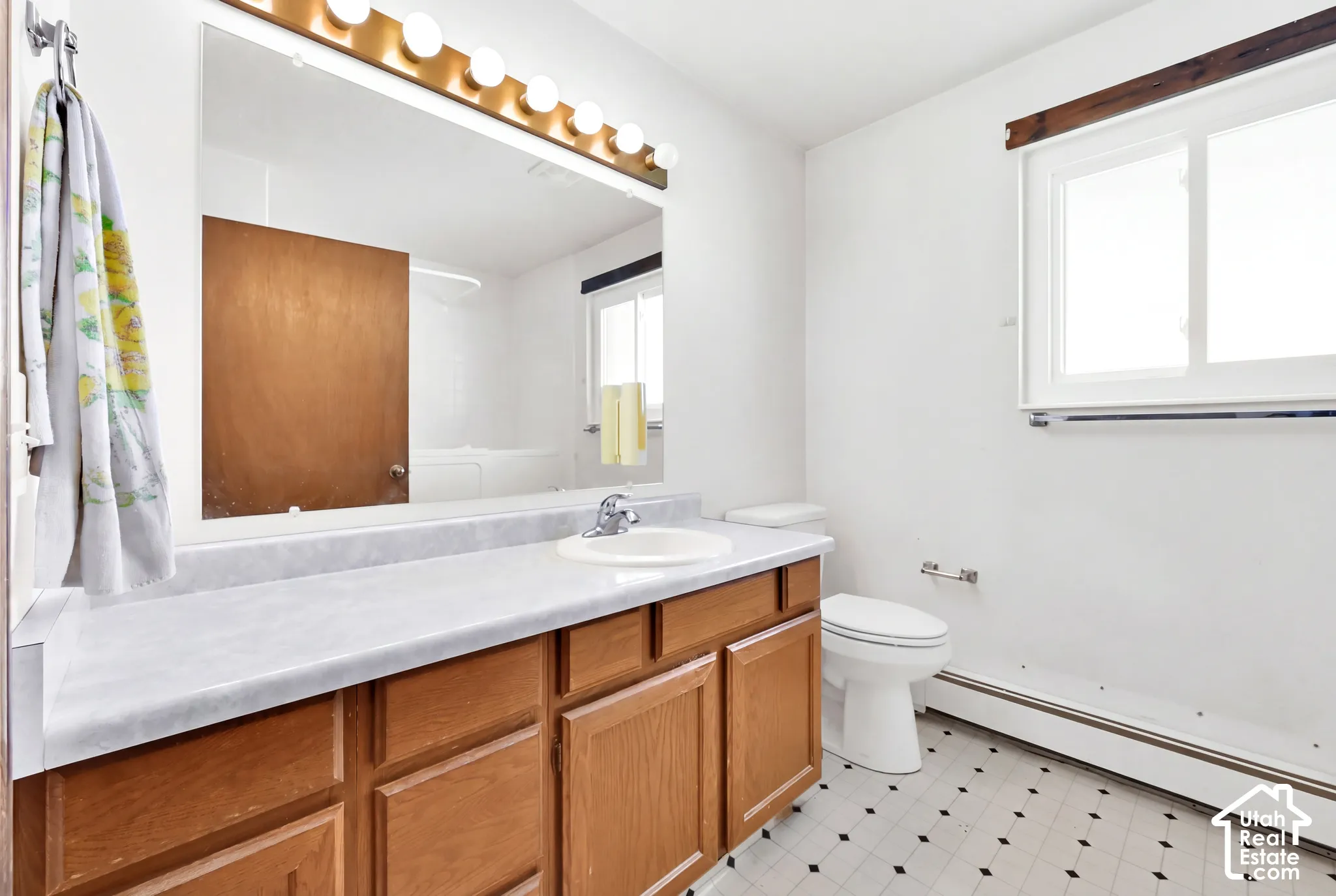 Bathroom featuring a vanity with wood-finish cabinetry, a large wall-mounted mirror, overhead vanity lighting, a window, and patterned floor tiling