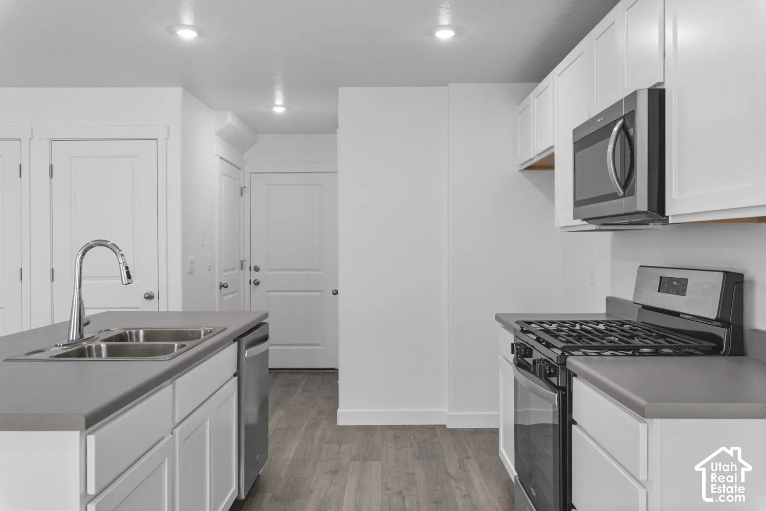 White shaker cabinetry, stainless steel appliances, wood-finish flooring, recessed lighting, and a kitchen island with a double basin sink