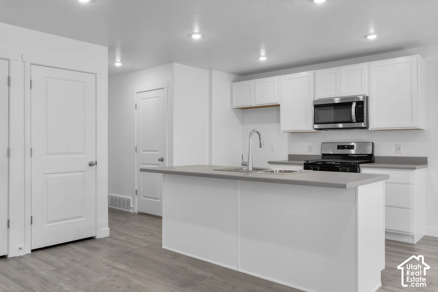 Kitchen featuring white cabinetry, grey countertops, stainless steel appliances, a center island with an undermount sink, and wood-finish flooring