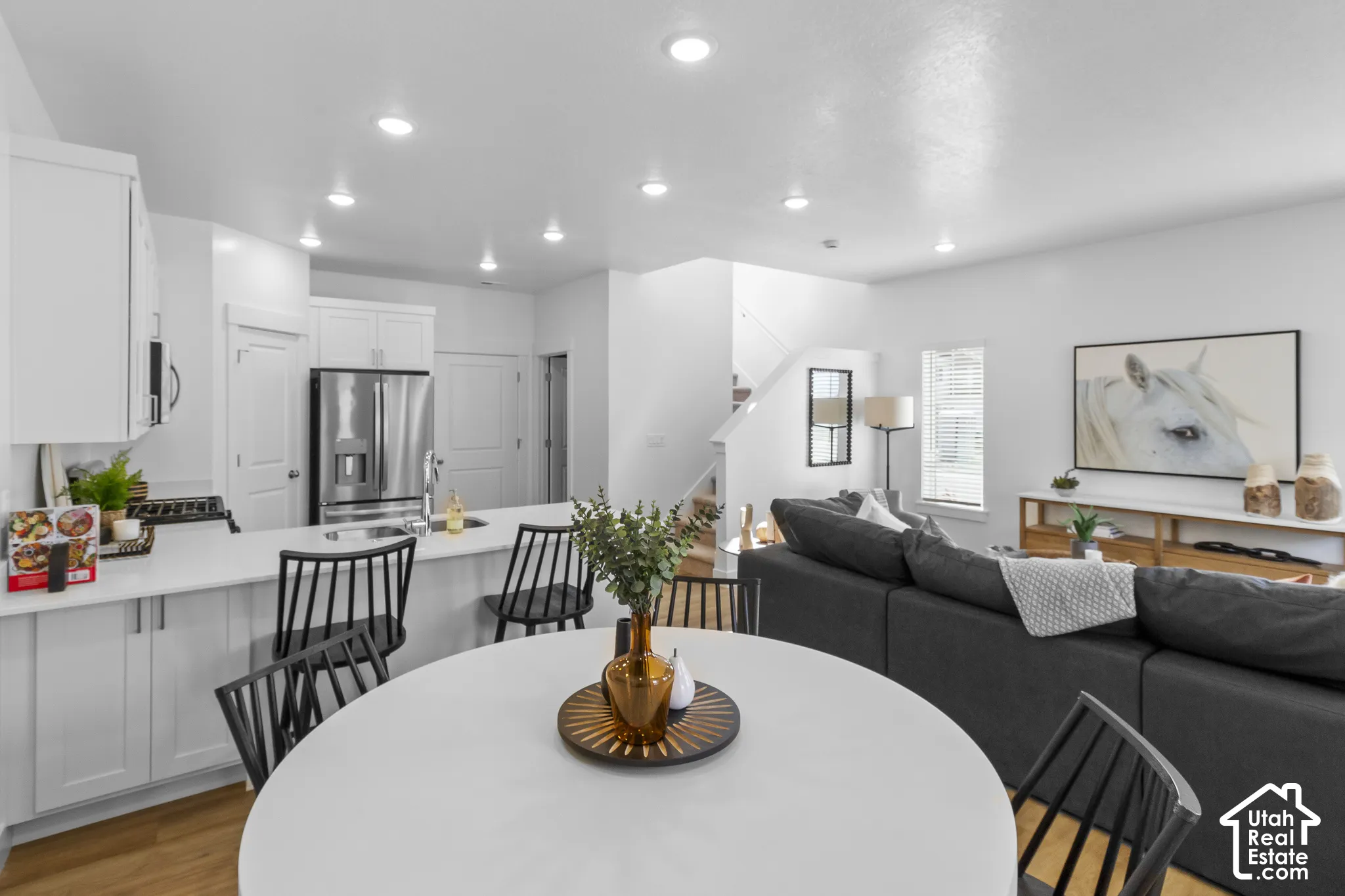 Open-concept living space featuring a modern kitchen with white cabinetry and stainless steel appliances, a kitchen island with seating, recessed lighting throughout, and wood-finish flooring