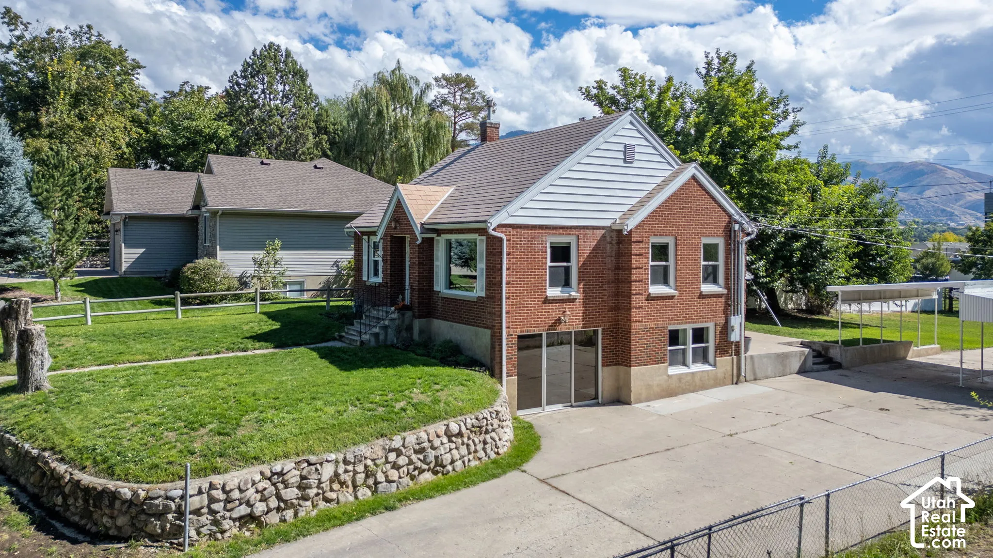 Red brick exterior with white gable siding, established landscaping, stone retaining wall, and a paved drive