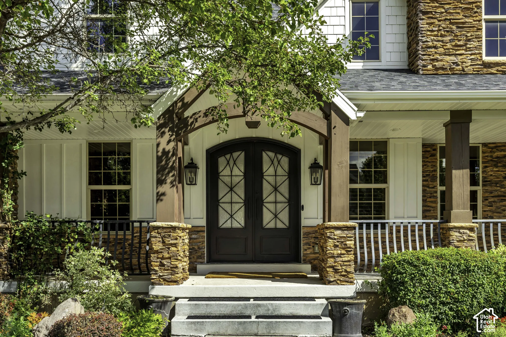 Arch-top double entry doors with decorative glass, stone veneer accents, board and batten siding, multiple windows with grilles, and a covered porch