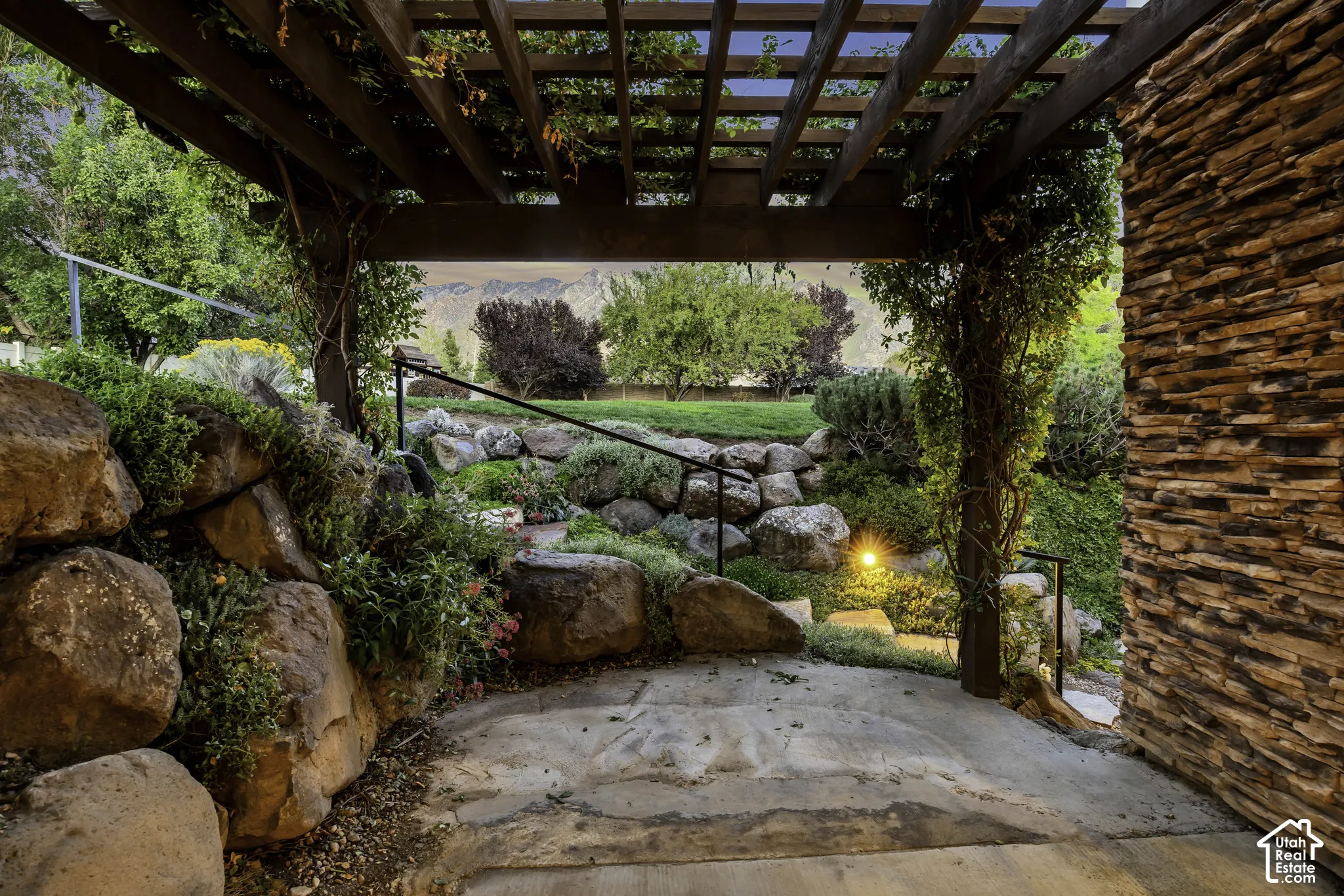 Wood pergola structure with climbing greenery, natural stone retaining walls, metal handrails, integrated landscape lighting, and a stacked stone exterior wall