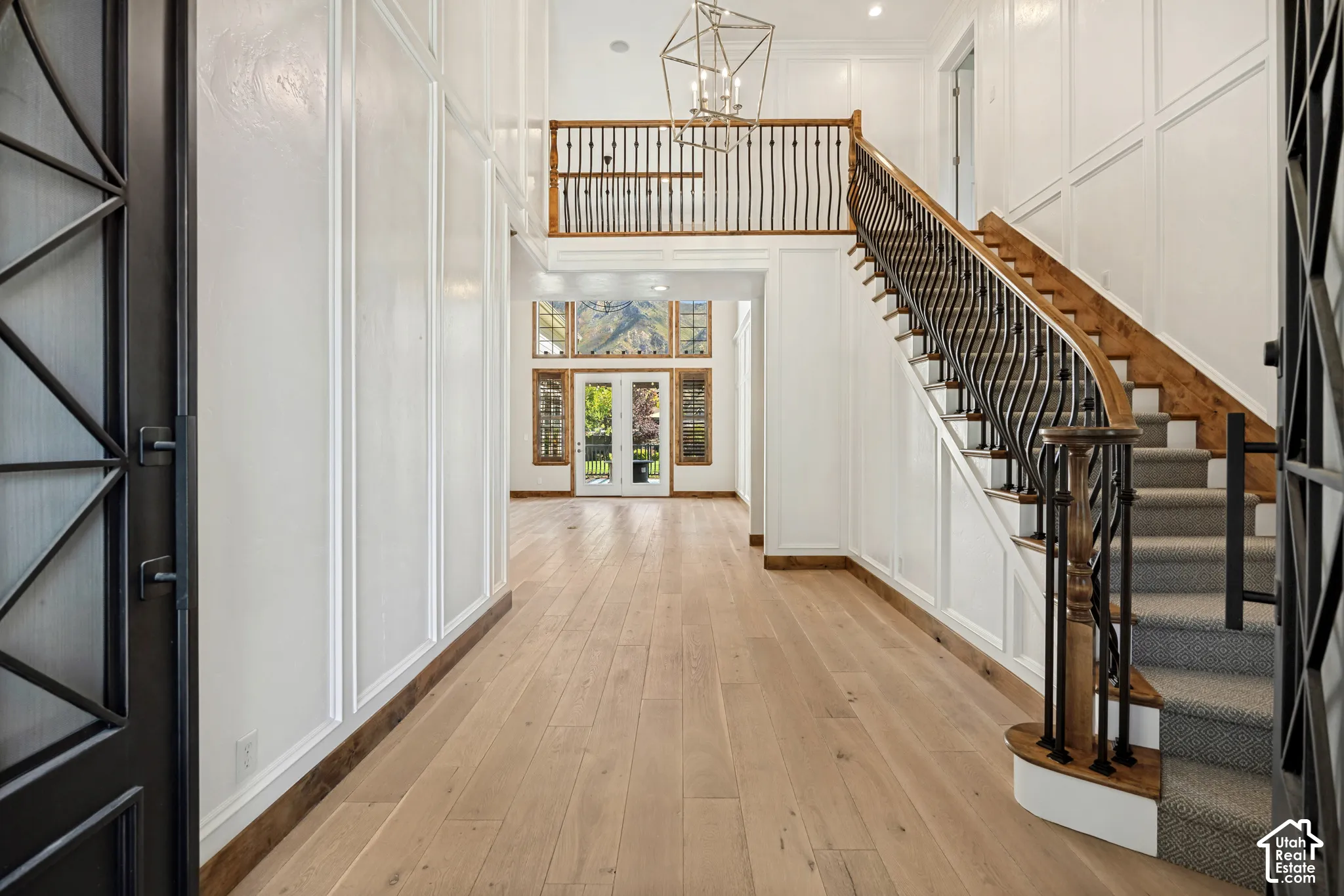 Two-story entryway featuring a grand staircase with wrought iron balusters, wood-finish flooring, white paneled walls, and a decorative chandelier