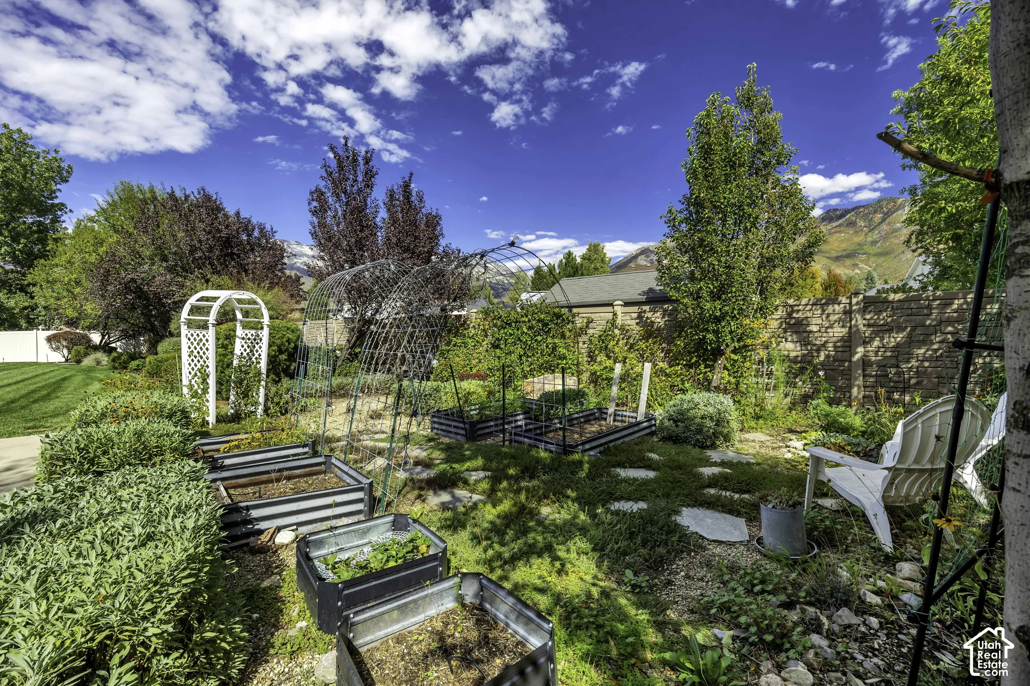Expansive garden area featuring multiple raised beds, a white garden trellis, stepping stone pathways, and mature privacy trees