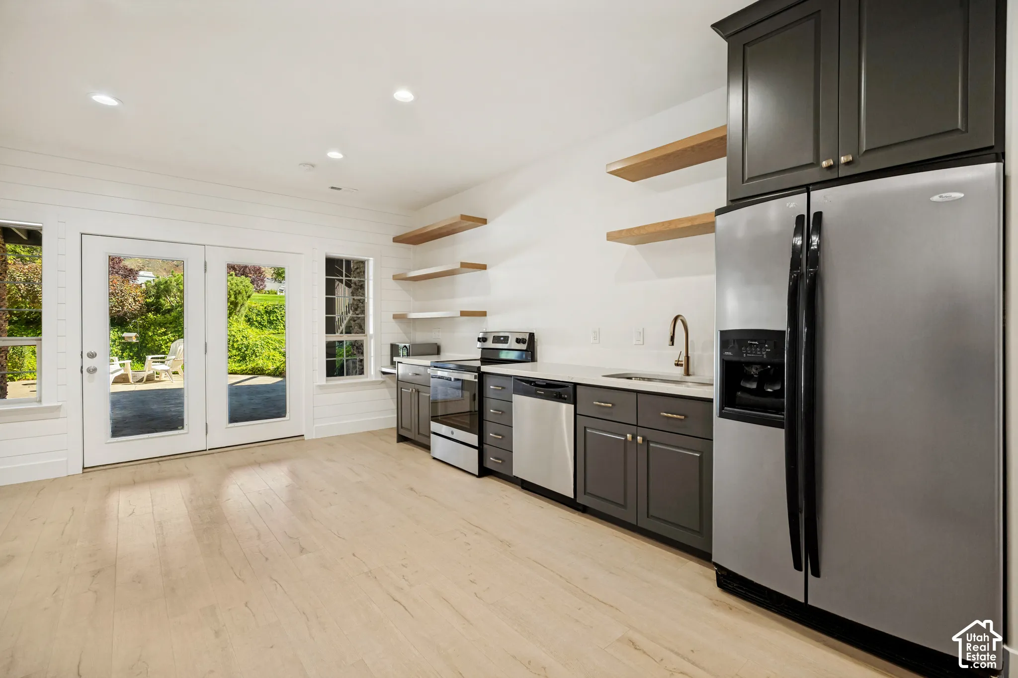 Light wood-finish flooring, white shiplap wall accents, stainless steel appliances, dark gray cabinetry, and floating wood shelves