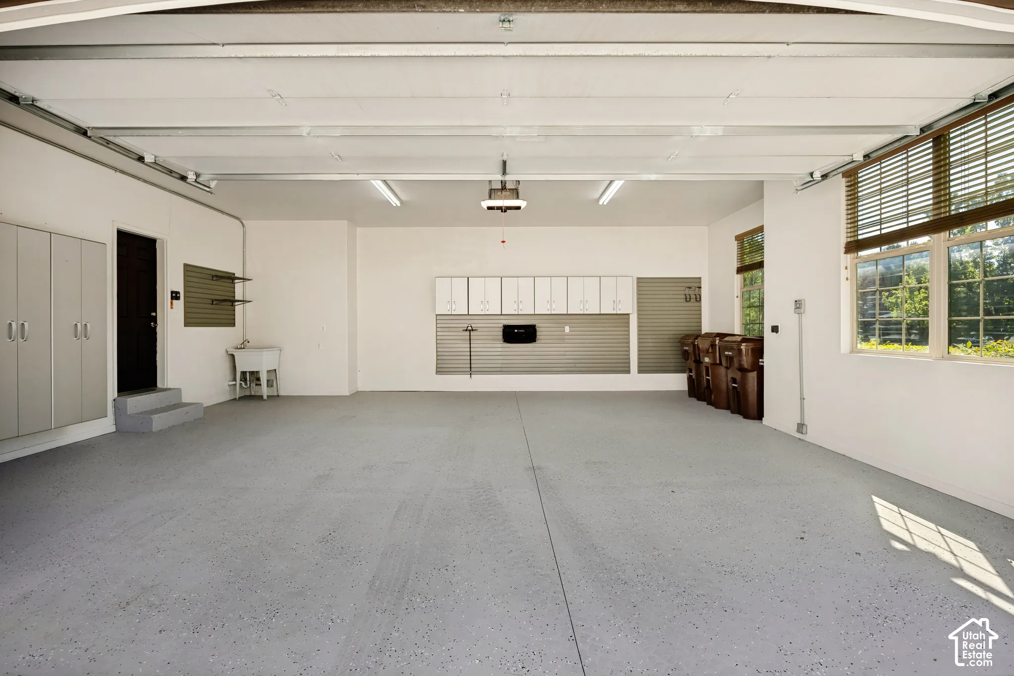 Expansive garage with durable epoxy flooring, multiple windows featuring wood-finish blinds, abundant white cabinetry, and a utility sink