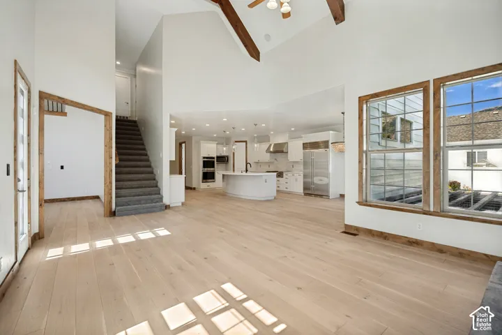 Vaulted ceiling with exposed wood beams, wood-finish flooring, white cabinetry, kitchen island with sink, and double windows