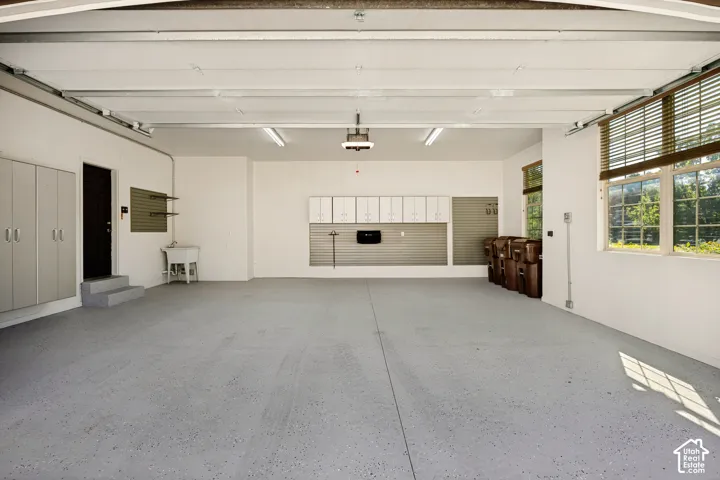 Expansive garage with durable epoxy flooring, multiple windows featuring wood-finish blinds, abundant white cabinetry, and a utility sink