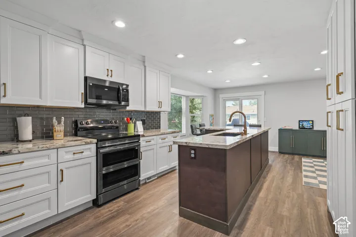 White cabinetry with gold-tone hardware, dark stainless steel appliances, grey subway tile backsplash, light-toned countertops, and a contrasting dark brown kitchen island