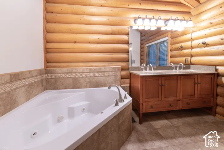 Log wall construction, corner soaking tub with deck-mounted fixtures, dual sink vanity with wood cabinetry, and a large vanity mirror with overhead lighting