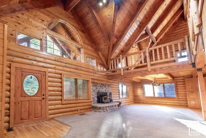 Expansive living area with log construction, soaring vaulted ceilings, an upper-level loft with log railing, a stone fireplace, and abundant natural light through multiple windows