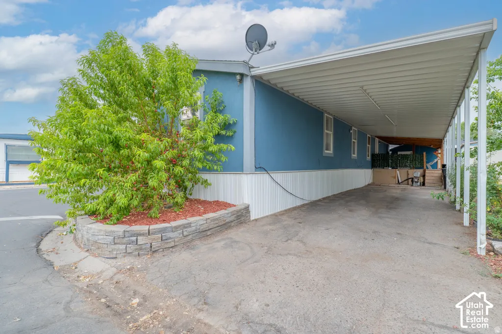 Covered carport extending the length of the home, blue siding, white trim, and a decorative stone-faced planter with established landscaping
