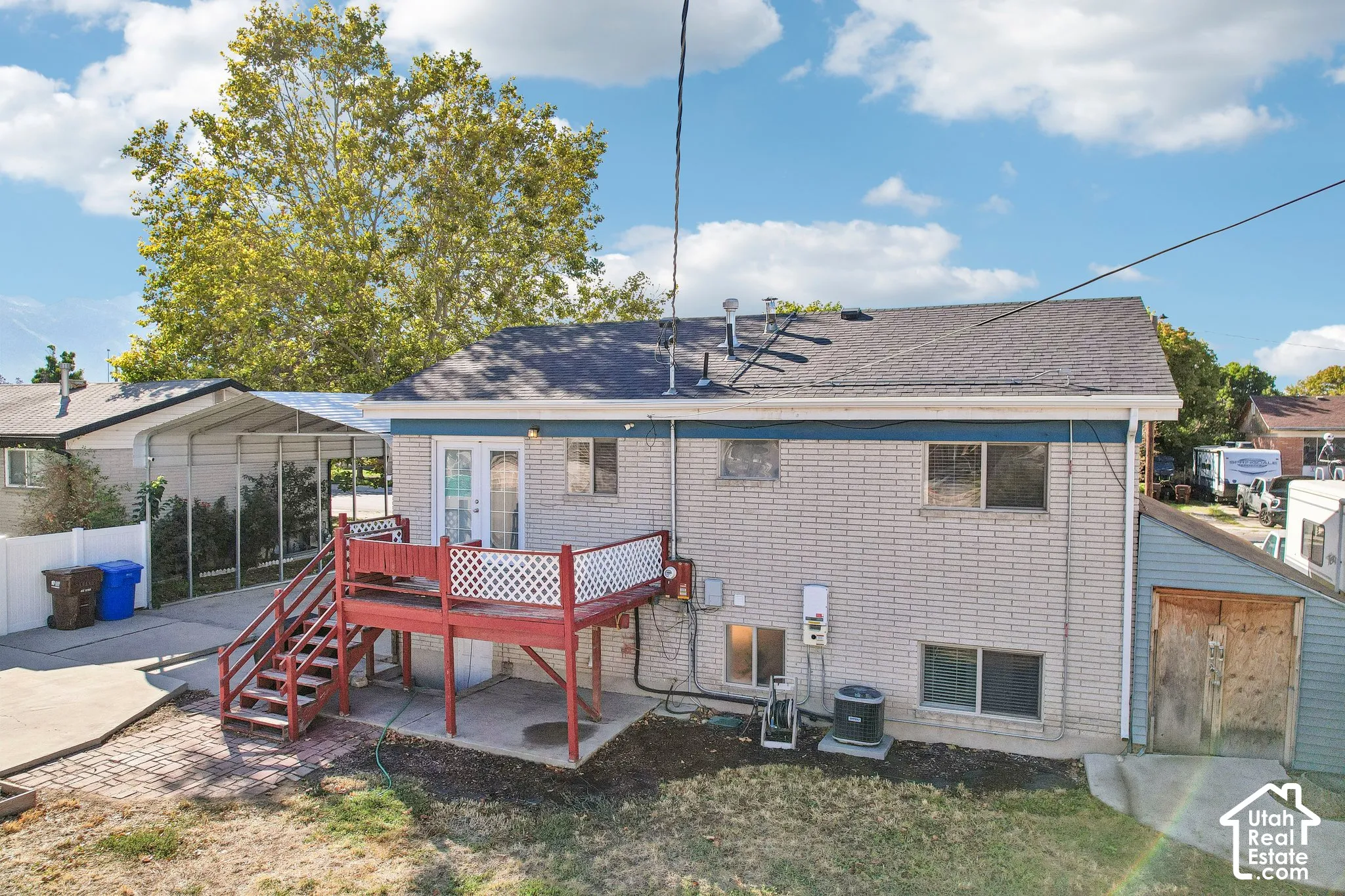 Rear exterior featuring a red-stained wooden deck with white lattice railing, light-colored brick siding, a ground-level patio, and an attached carport