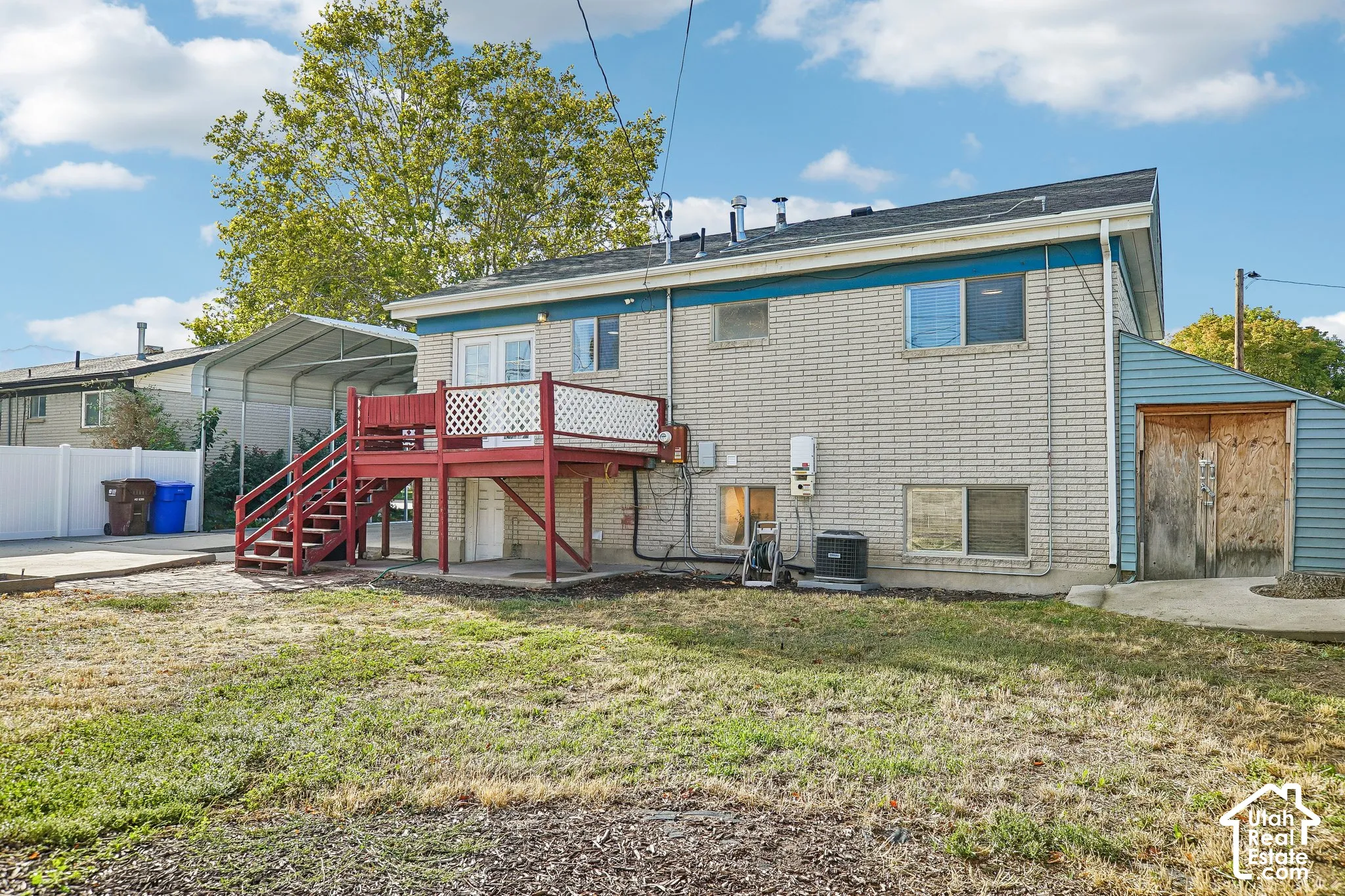 Rear exterior featuring a red deck with lattice railing, light-colored brick siding, a blue-sided attached shed, a white privacy fence, and a metal carport structure