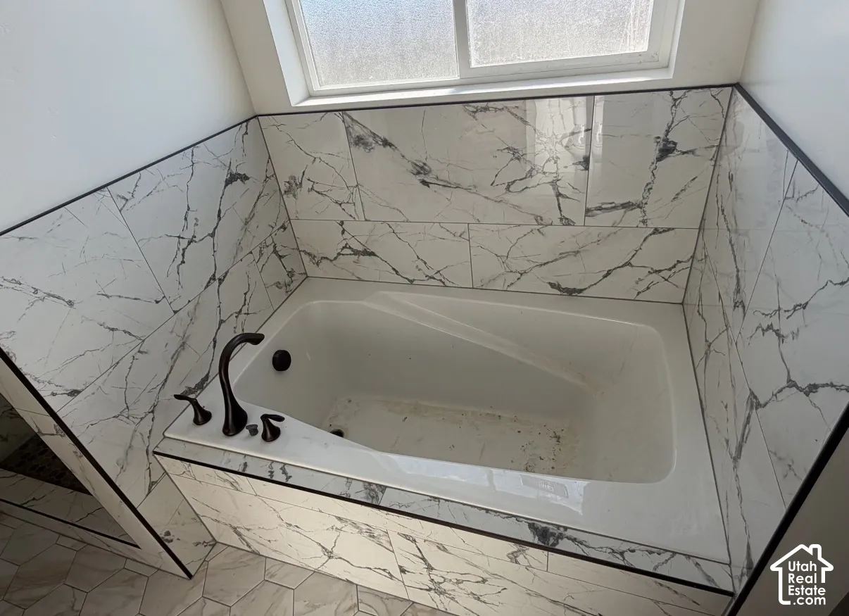 White soaking tub with dark bronze fixtures, surrounded by white and gray patterned tile, featuring a privacy window