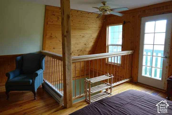 Natural wood flooring, wood paneling, wood railing with metal spindles, ceiling fan, and white-framed windows and door