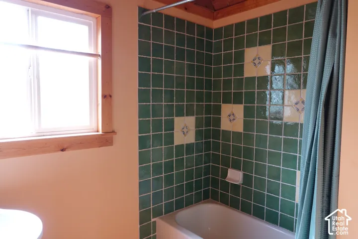 Tub-shower combination featuring green and patterned accent wall tile, wood-paneled ceiling, and a window with wood trim