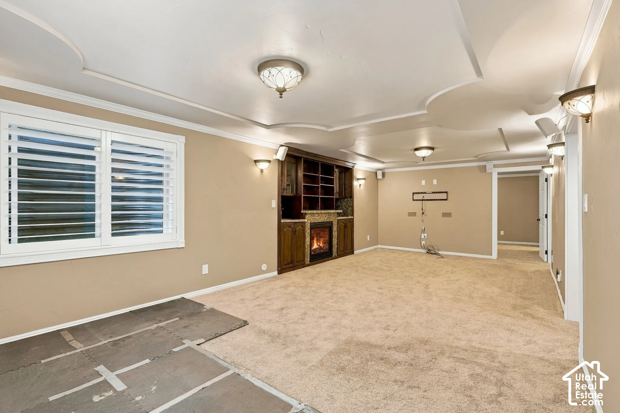 Recessed ceiling with decorative molding, built-in media cabinetry, fireplace, wall sconces, and plantation shutters