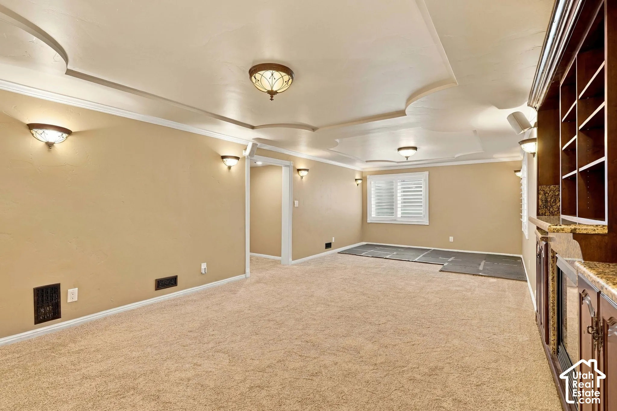 Tray ceiling with decorative moldings, built-in cabinetry with display shelving, window with louvered shutters, wall sconce lighting, and plush carpeting