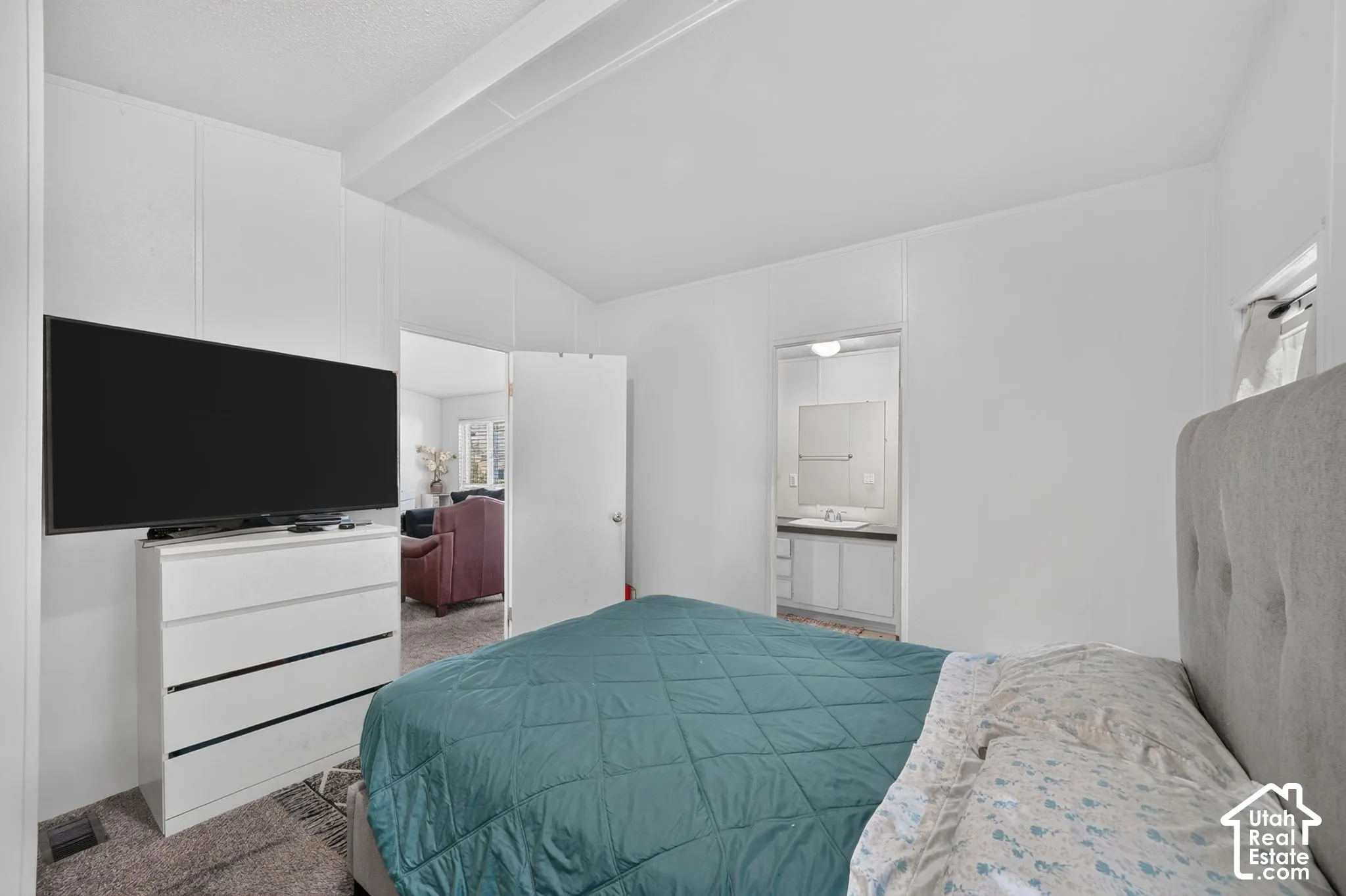 White paneled walls with a vaulted ceiling, gray carpeting, and an open doorway revealing a bathroom with a vanity and mirror