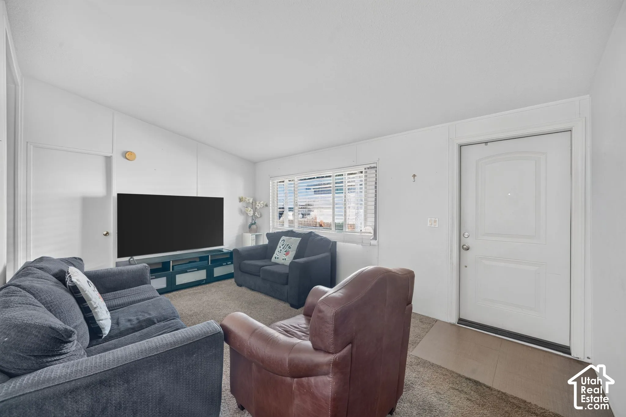 Vaulted ceiling, white painted walls, window with blinds, and white entry door